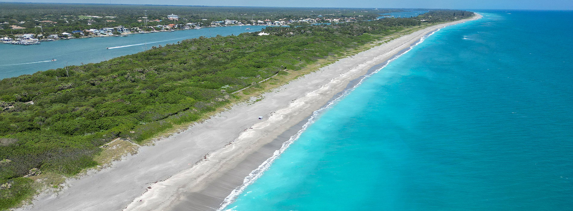 Aerial view of a sandy beach with clear blue water, adjacent to a forested area and a coastal cityscape.