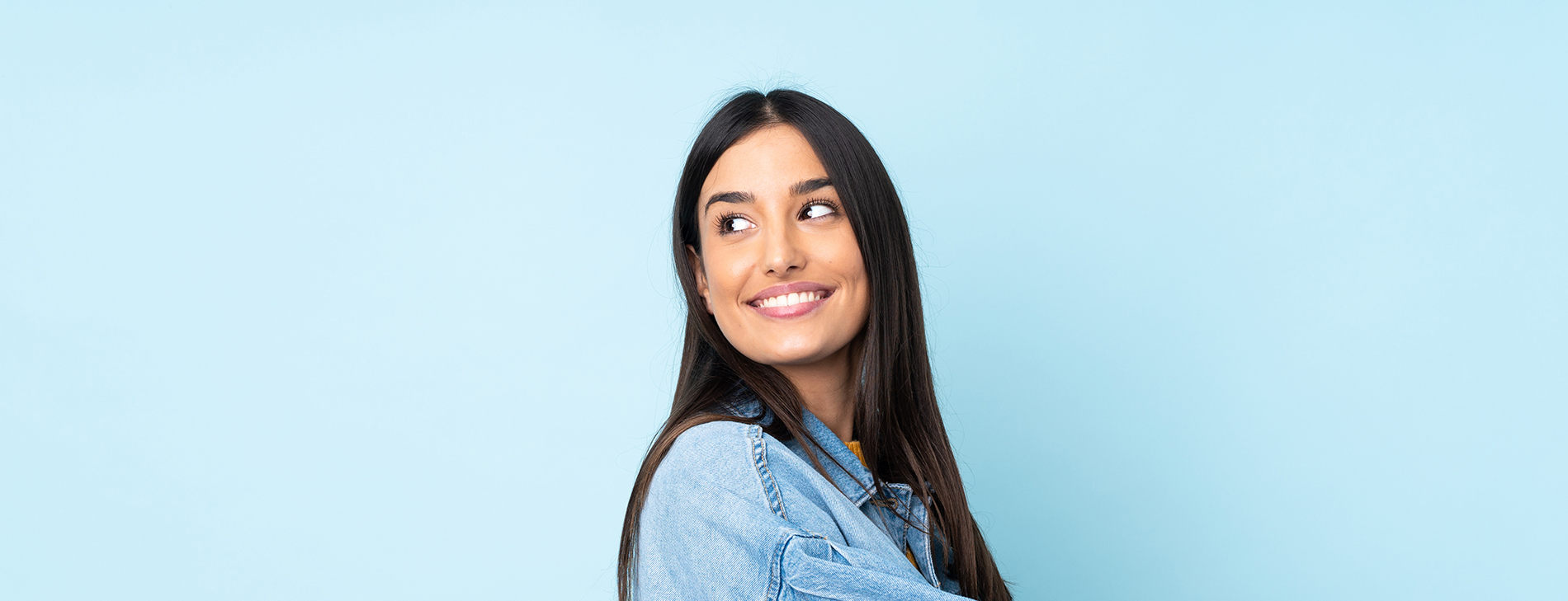 The image is a photograph of a woman with light skin, smiling at the camera. She appears to be in her late twenties or early thirties and has long hair. Her eyes are looking directly at the camera, and she is holding up her index finger near her mouth as if she s making a point or emphasizing something. The background is plain and light-colored, which suggests that this could be a stock photo used for various purposes such as advertising, personal branding, or lifestyle content.