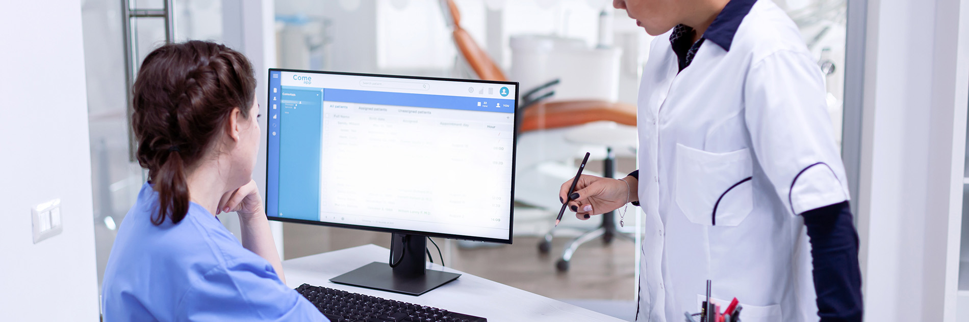 A healthcare professional in a white coat and stethoscope standing at a desk with a computer monitor, interacting with a patient seated in front of the screen.