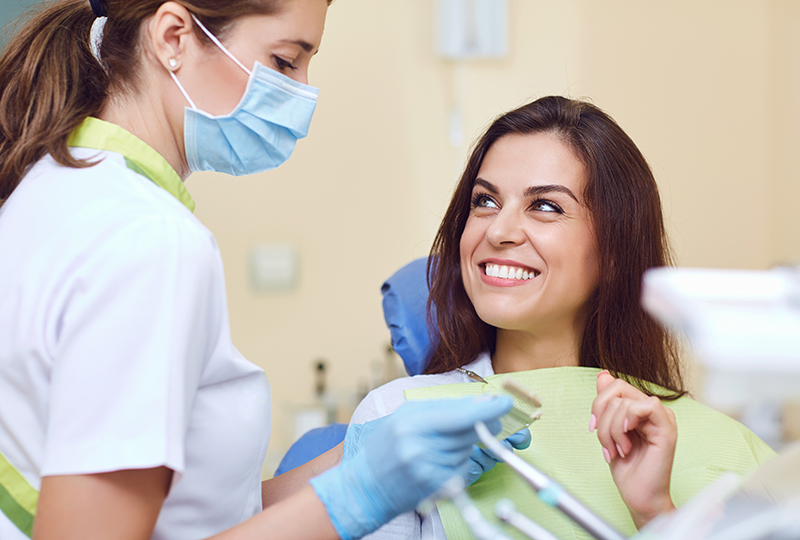 The image shows a dental hygienist in a white coat and mask, smiling at the camera, while a woman with a blue apron is seated behind her.