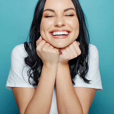 The image features a person with dark hair, wearing a white top, smiling broadly and covering their mouth with both hands.