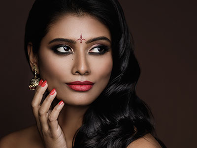 The image is a close-up portrait of a woman with dark hair and makeup, wearing red nail polish.